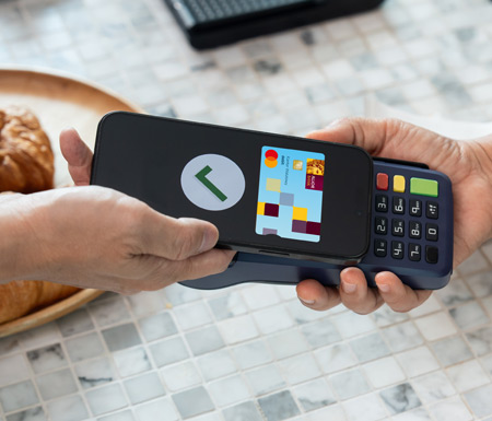 Ręka trzymająca smartfon z otwartym  portfelem cyfrowym. W portfelu widoczna karta Kantoru Walutowego izielony znacznik potwierdzający udaną transakcję. W tle widać terminal płatniczy.